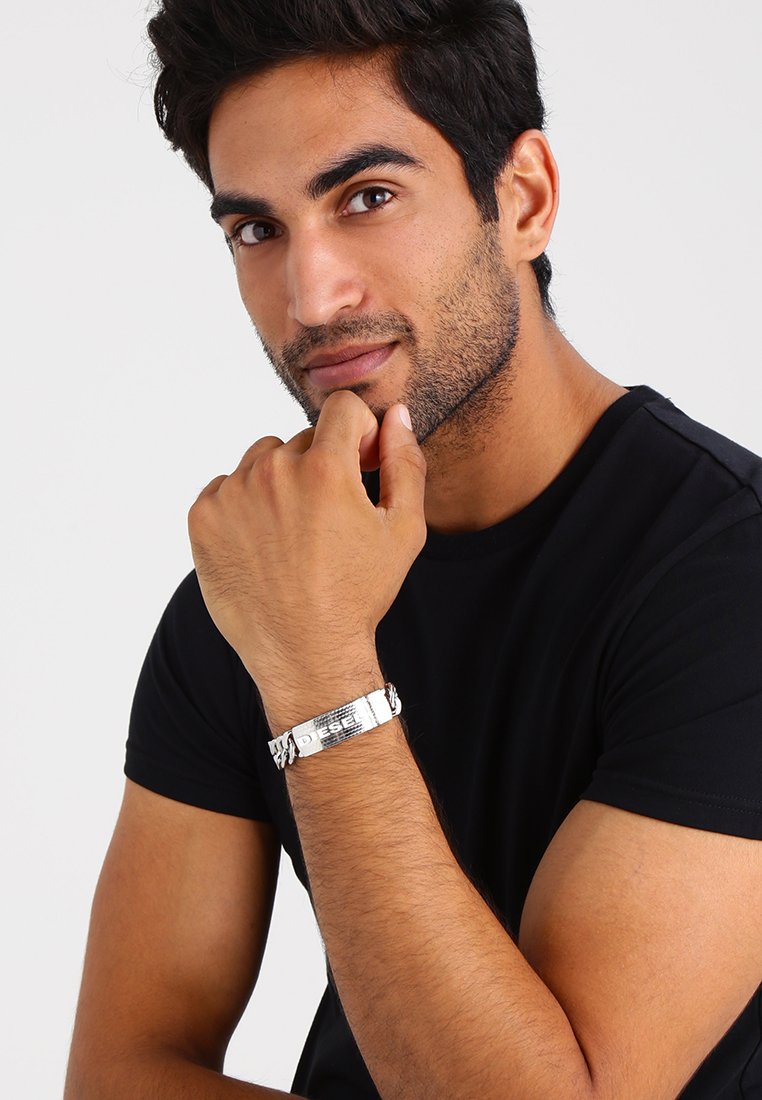 Bracelet en argent avec un bandeau texturé et un accent gravé affichant "DIESEL." Le modèle porte un t-shirt noir et repose sa main sur son menton.