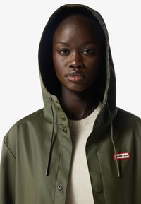 Mujer joven que lleva un impermeable verde oliva con capucha puesta y un suéter crema, mirando al frente con expresión neutra.