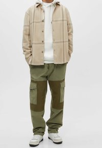 Personne portant une surchemise à carreaux beige par-dessus un sweat à capuche blanc, un pantalon cargo bicolore vert olive, et des baskets blanches, debout les mains dans les poches.