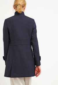 Abito blu navy, sartoriale, realizzato in tessuto liscio con un colletto alto, cuciture strutturate e polsini con bottoni. Presenta una vita cinte e un retro svasato.