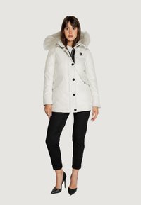 Parka bianca isolata con cappuccio foderato in pelliccia sintetica, bottoni neri e tasche laterali, abbinata a pantaloni neri aderenti e scarpe con tacco alto.