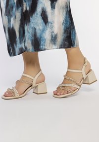 Sandales à talons beiges avec plusieurs lanières ornées d'accent en cristal, un talon carré et une texture en cuir lisse.
