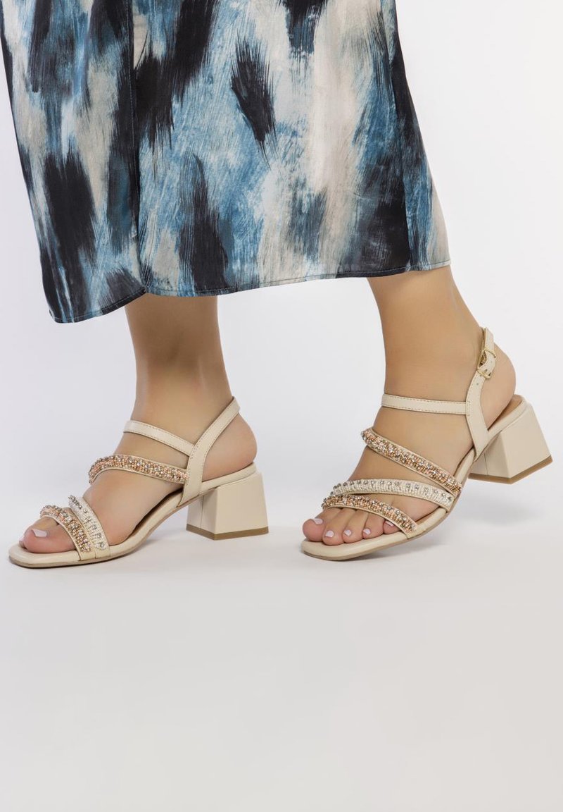 Sandales à talons beiges avec plusieurs lanières ornées d'accent en cristal, un talon carré et une texture en cuir lisse.