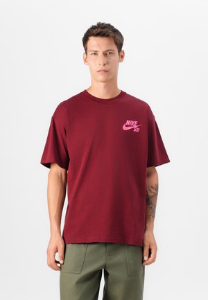 Bordo pamučna majica s ružičastim Nike SB logotipom na lijevoj strani prsiju. Dizajn uključuje kratke rukave i klasični okrugli izrez.
