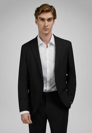 Młody mężczyzna ubrany w czarny garnitur i białą koszulę stojący z jedną ręką w kieszeni na jednolitym szarym tle.