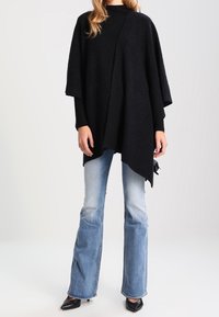 Svart poncho med lös passform, tillverkad av mjukt material. Har korta ärmar och en v-ringad design, i kombination med utsvängda ljusblå jeans.