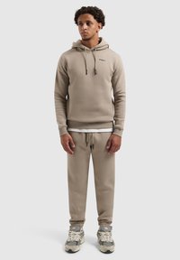 Jongen staat in een beige hoodie, bijpassende joggingbroek, wit onderhemd en grijs-witte sneakers tegen een effen achtergrond.