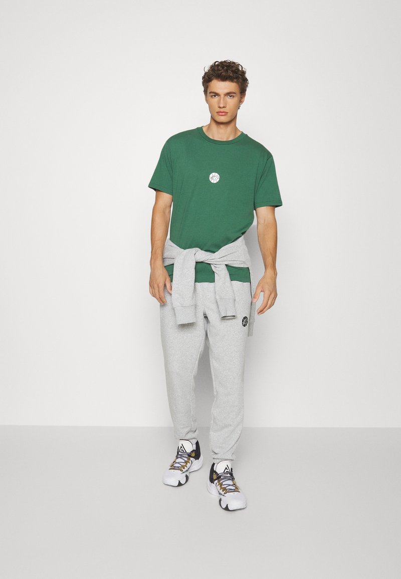 Grünes Baumwoll-T-Shirt mit einem zentralen Logo, graue Jogginghosen mit elastischem Bund und weiße Sneakers mit schwarzen Akzenten.