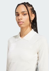 White long-sleeve polo shirt with a collar, featuring subtle Adidas branding and logo. Smooth fabric with a slight sheen.