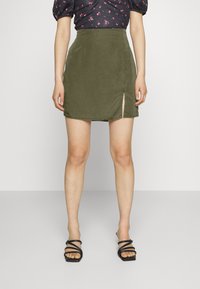 Saia mini em camurça verde-azeitona com uma fenda lateral, apresentando um design ajustado e uma textura suave. Combinada com sandálias de salto alto pretas com tiras.