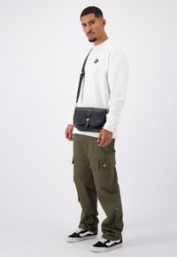 Zwarte leren schoudertas met flapsluiting, gedragen over een witte lange mouwen shirt en olijfgroene cargobroek, met zakken en een logo.