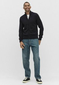 Granatowy kardigan zapinany na zamek, wykonany z dzianiny, w połączeniu z jasnoniebieskimi dżinsami i czarnymi sneakersami, o luźnym kroju i ściągaczach na rękawach.