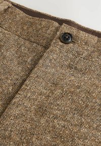 Țesătură de tweed maro cu o țesătură texturată, având un nasture negru și un brâu maro. Culoarea generală prezintă variații subtil.