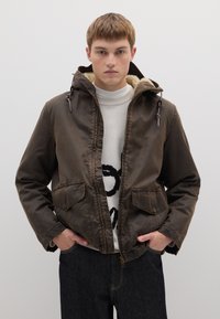 Braune, strukturierte Jacke mit einem mit Pelz gefütterten Kapuzen, Kordelzügen und zwei Vordertaschen. Über einem cremefarbenen Pullover getragen. Dunkle Jeans vervollständigen den Look.