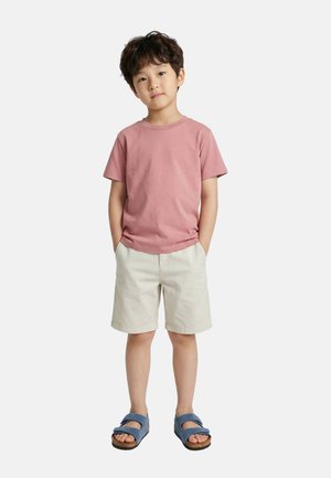 Un jeune garçon portant une chemise rose à manches courtes, un short beige et des sandales bleues, debout les mains dans les poches devant un fond uni.