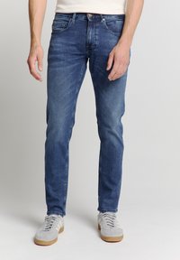 Donkerblauwe, slim-fit jeans van denim. Bevat voorzakken, riemlussen en subtiele vervaging. Gecombineerd met lichtgrijze sneakers.