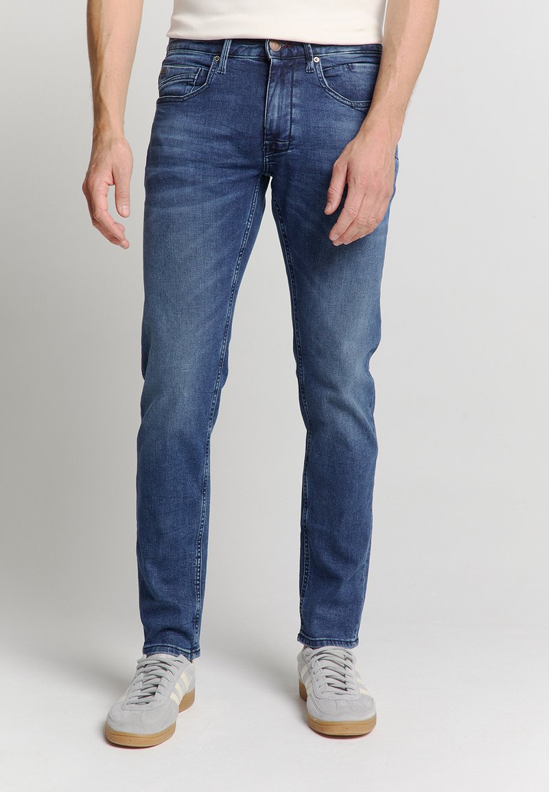 Donkerblauwe, slim-fit jeans van denim. Bevat voorzakken, riemlussen en subtiele vervaging. Gecombineerd met lichtgrijze sneakers.