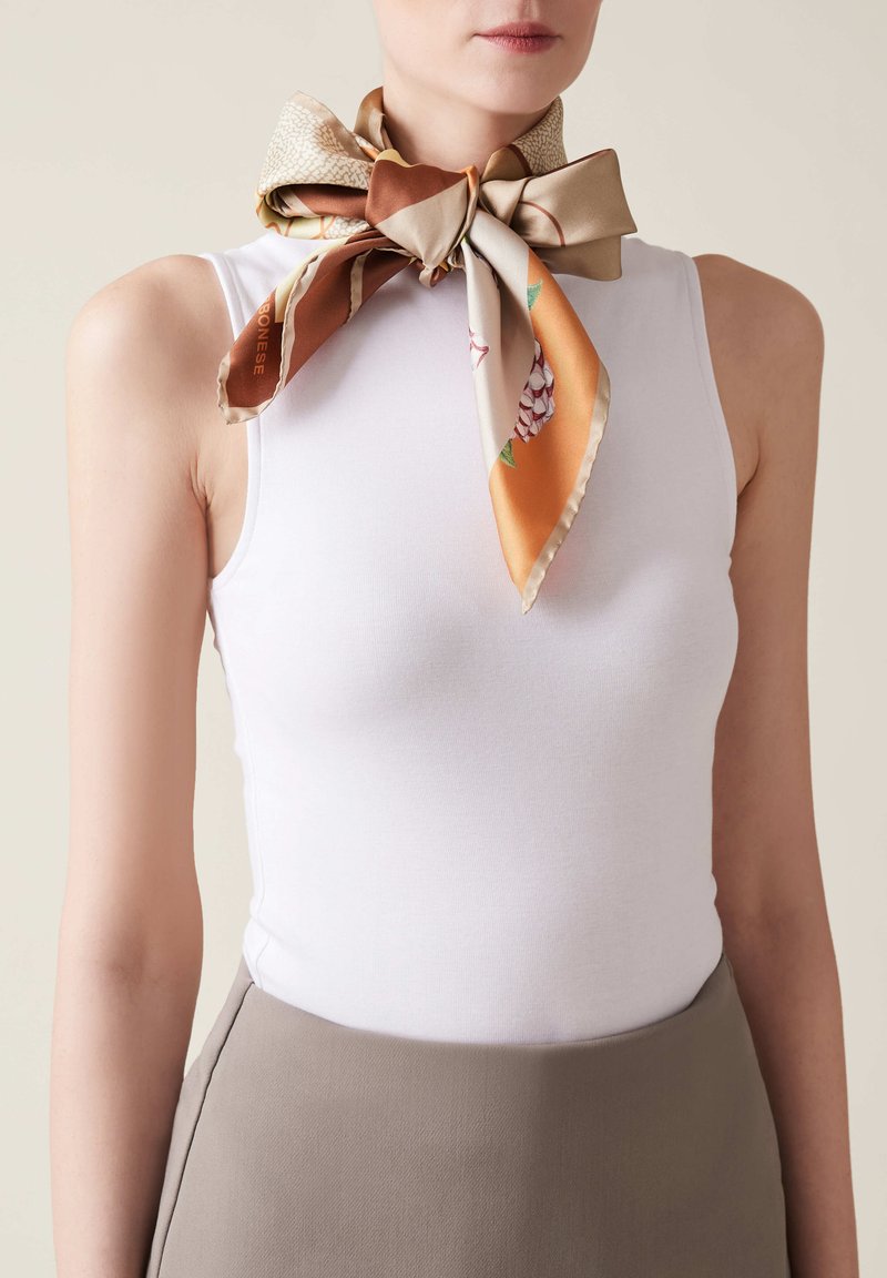 Top in cotone bianco aderente abbinato a una sciarpa di seta patterned legata a fiocco attorno al collo, con tonalità di beige, marrone e arancione.