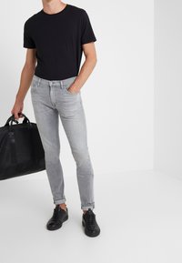 Černé triko s kulatým výstřihem, světlé šedé slim-fit džíny, matné černé tenisky a černá kožená taška přes rameno, vše na jednobarevném bílém pozad�í.