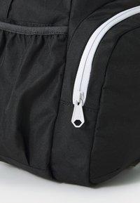 Mochila preta com um material texturizado, fecho éclair branco em destaque com um puxador em formato hexagonal e um bolso lateral com tecido franzido.