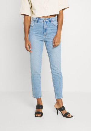 Persoon die lichtblauwe hoogwaterjeans, een beige cropped shirt, zwarte sandalen met hak en gouden ringen draagt, staand tegen een eenvoudige lichte achtergrond.