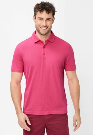 Lächelnder Mann mit lockigem Haar in einem knallpinken kurzärmligen Poloshirt und dunkelrosa Hose, stehend vor weißem Hintergrund.