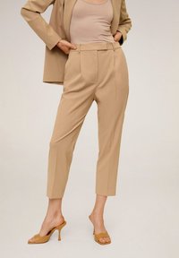 Femme portant un pantalon beige ajusté, des talons assortis et un blazer beige avec un débardeur beige clair sur un fond uni.