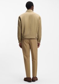 Bēša satīna bombardiera jaka ar tīru dizainu, kas aprīkota ar muguras klāju, sānu kabatām un gludu tekstūru. Kombinēta ar šaurām bēšām biksēm.