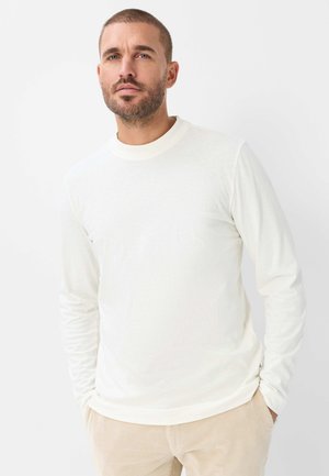 Mand med kort hår og skæg iført en simpel hvid langærmet skjorte og beige bukser, der står foran en hvid baggrund.