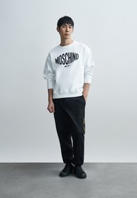 Άσπρο βαμβακερό φούτερ με μαύρο κείμενο "MOSCHINO" και λεπτομέρεια "since 1983", συνδυασμένο με μαύρο παντελόνι που διαθέτει τσέπη με φερμουάρ. Μαύρα αθλητικά παπούτσια.