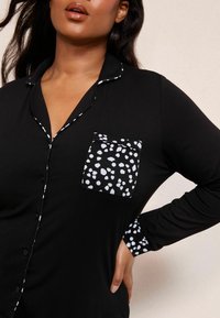 Cămașă de pijama neagră cu mâneci lungi, cu detalii de piping alb, un buzunar alb cu pete și o textură de material neted. Design cu închidere cu butoane.