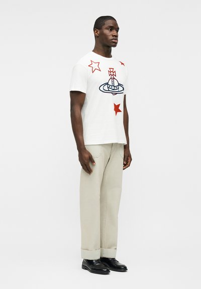 Άνδρας όρθιος, φορώντας λευκό μπλουζάκι με κόκκινα αστέρια και μπλε έμβλημα σε σχήμα σφαίρας, μπεζ παντελόνι με ρεβέρ και μαύρα επίσημα παπούτσια, μπροστά σε απλό φόντο.