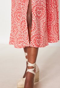 Onderbenen van een persoon die beige wedge espadrilles met enkelbanden draagt, samen met een midi-jurk met een rood en wit patroon en een split aan de voorkant.