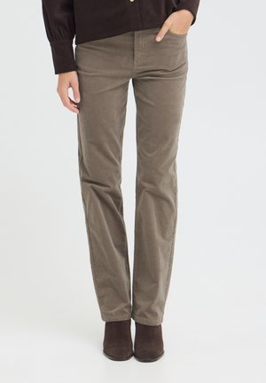 Personne portant un pantalon slim marron, une chemise marron foncé et des bottines marron foncé, debout avec une main dans la poche devant un fond uni.