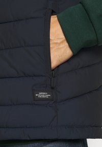 Granatowa pikowana kamizelka puchowa z kieszenią na suwak i oznaczoną metką. Teksturowana tkanina, pikowany wzór oraz widoczny zielony mankiet pod spodem.