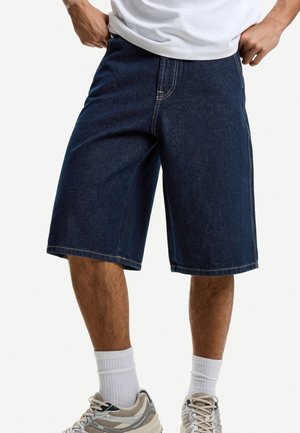 Adult wearing dark blue denim knee-length shorts, white socks, and beige sneakers, with hands resting on waistband.