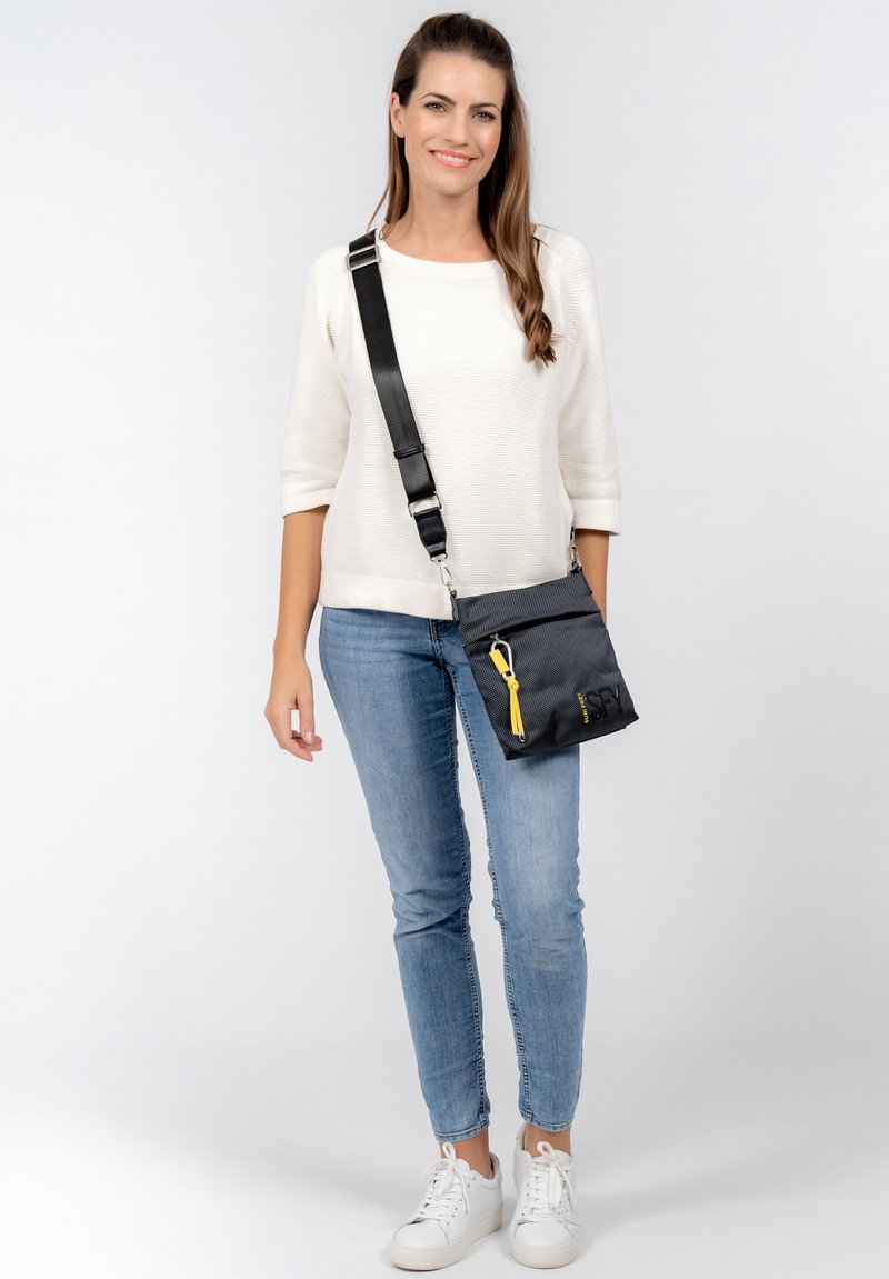 Borsa a tracolla nera con tessuto testurizzato, zip gialla e tracolla regolabile. Il modello indossa un maglione bianco e jeans blu.