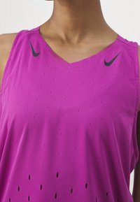 Bezrekawnik sportowy z dekoltem w serek, wykonany z jasnoróżowej tkaniny z perforowanym wzorem i czarnym logo Nike na lewym ramieniu.