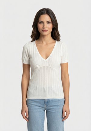 Femme aux cheveux noirs mi-longs portant un haut blanc en maille à manches courtes et col en V, et un jean bleu clair, debout devant un fond uni.