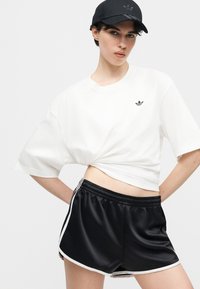 Άτομο που φοράει λευκό κοντό t-shirt Adidas, μαύρο αθλητικό σορτς Adidas με λευκές ρίγες και μαύρο καπέλο Adidas, μπροστά σε απλό φόντο.