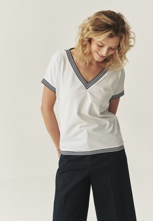 T-shirt blanc à col en V avec des bordures rayées bleu marine autour du col et des manches, fabriqué en tissu doux et lisse. Porté avec un pantalon sombre.
