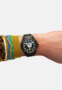 Schwarze Uhr mit gelben Akzenten, auffälligen Ziffern, strukturierter Zifferblatt und silbernen Details am Handgelenk. Farbige gestrickte Pulloverbündchen sichtbar.