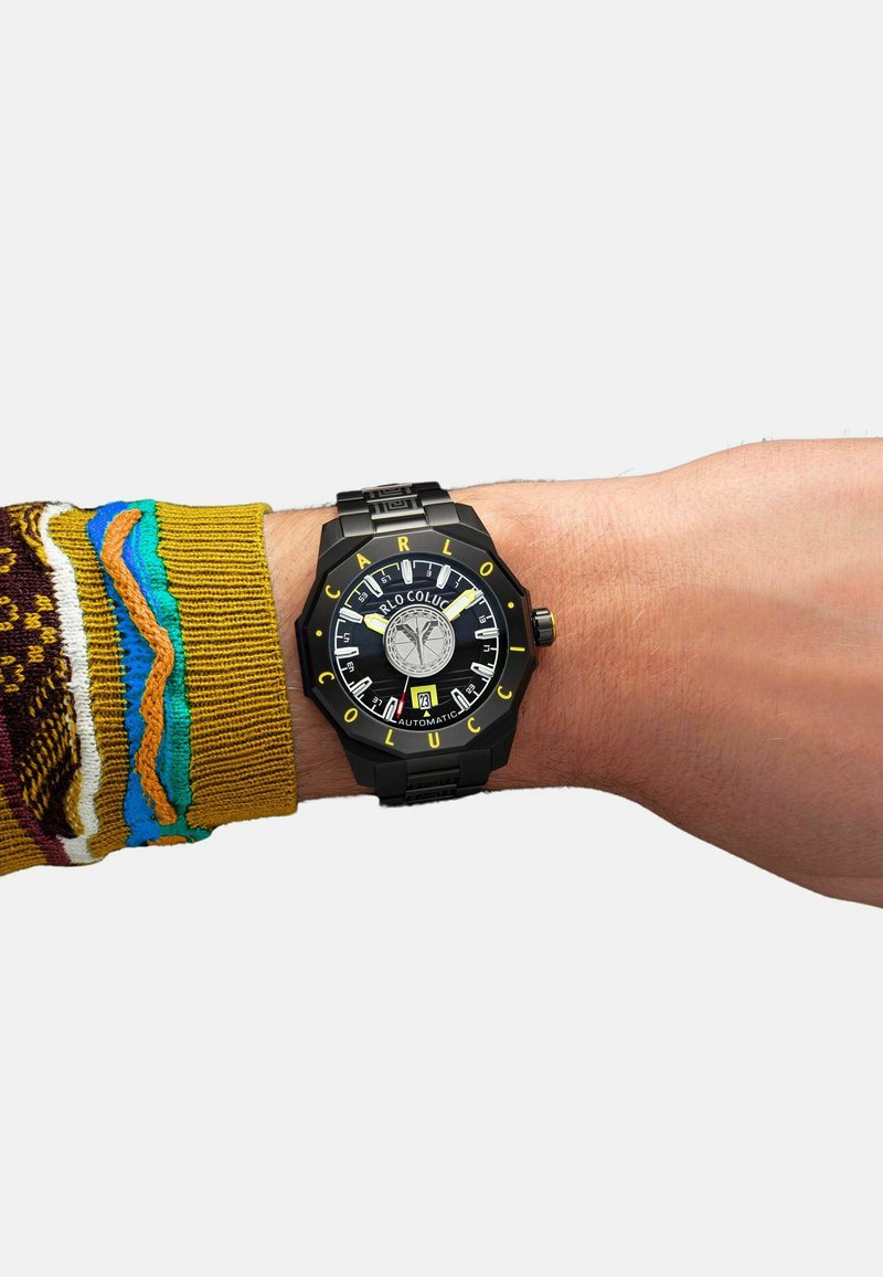 Schwarze Uhr mit gelben Akzenten, auffälligen Ziffern, strukturierter Zifferblatt und silbernen Details am Handgelenk. Farbige gestrickte Pulloverbündchen sichtbar.