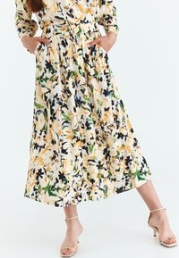 Rochie cu model floral, cu talie ajustabilă prin �șnur, confecționată dintr-un material ușor. Culorile includ galben, verde și negru pe un fundal crem.