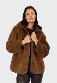 Braune Fleecejacke mit Kapuze, mit einem Reißverschluss vorne, kontrastierenden dunklen Details und weit geschnittenen Ärmeln. Weicher Stoff mit lockerer Passform.