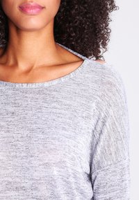 Haut gris clair, ample, avec un col large et un motif en maille texturé. Présente des découpes au niveau des épaules.