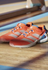 Coppia di scarpe da ginnastica arancioni con strisce bianche e accenti blu che riposano su un pavimento in legno vicino a una linea del campo da palestra.