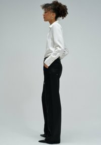 Camisa blanca de manga larga con puños abotonados, combinada con pantalones negros de pierna ancha con bolsillos laterales y un detalle de cadena. Zapatos negros de punta.