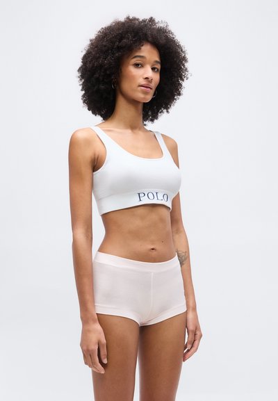 Γυναίκα με σγουρά μαλλιά, που φοράει ένα λευκό crop top με το κείμενο "POLO" και ανοιχτό ροζ εσώρουχο τύπου boyshort, στέκεται μπροστά σε ένα απλό φόντο.