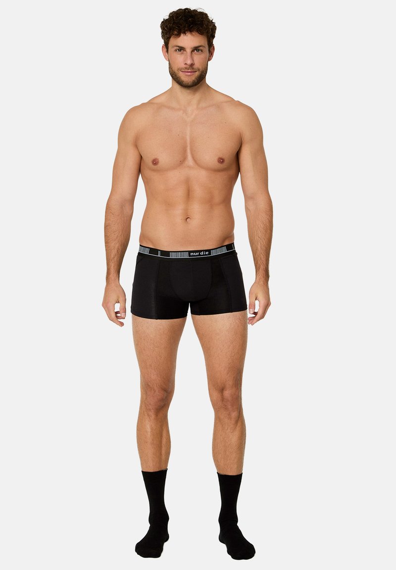 Vältränad man som står och bär svarta boxershorts och svarta strumpor, framför en enkel ljusgrå bakgrund.